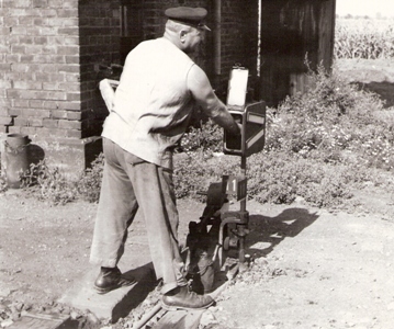  Výhybkár v pracovnom pri manipulácii s petrolejkou vo výhybk. lampe pri stavadle z režn. muriva, za ním kukurič, pole. Anon. cca 1960. 185 x 154 