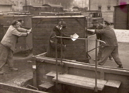  Skupina mužov v prac. so železn. čiapkami pretláča malý kontajner z rampy na nízkosten. vozeň s drev. kostrou na rampe nákl. obv. Bratislava hl. st. Anon., cca 1950. 172x125 