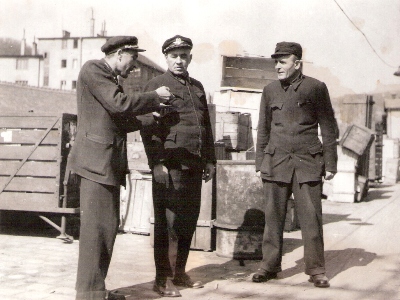  Traja muži v železn. rovnošatách na rampe skladu žst. Bratislava hl. st. Za nimi zložené sudy a debny. Anon., cca 1950. 145 x 110 