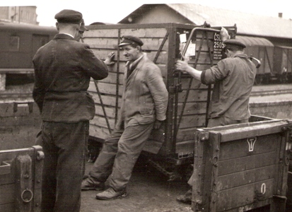  Štyria muži v pracovnom so železn. čiapkami pri nakladaní malého kontajneru na nízkostenný vozeň s drev. kostrou. Text: Žst. Bratislava hl. st. Anon., cca 1950. 170 x 125 