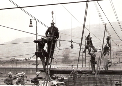  Rekonštrukcia trakč. vedenia v Žiline (zrejme). Robotníci v trolejovej inštalácii, osvetľ. stožiare, sklad, vzadu kopec.Foto: J. Hauzer, cca 1977. 173 x 118 
