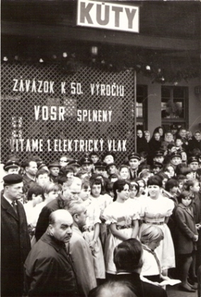  Zástup ľudí v žel. rovnošatách a civile, dievčatá v krojoch na peróne žst. Kúty. Nápis: Záväzok k 50. Výročiu VOSR splnený, vítame 1. elektrický vlak. Anon., 1967. 160 x 238 