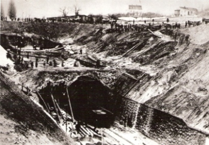 Bratislava hl. st. - tunel počas prestavby. Pohľad na stanič. tunelový otvor bez portálu zhora. Vpravo zárub. múr, všade pózujúci ľudia s náradím, káry so zeminou, drevené lešenie. Anon., cca 1900. 174 x 122 