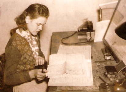  Žena v civile sediaca za pís. stolom. Na stole atrament, pís. potreby, razítka, telefón PE a selektor a nejaký denník. Anon., cca 1955. 115 x 85 
