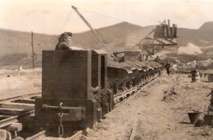  Motorový rušník s rušňovodičkou ťahá plechové huntíky ložené zeminou. V pozadí lanový báger. Text: Rekonštrukcia Trate družby. Anon., cca 1950. 167 x 110 