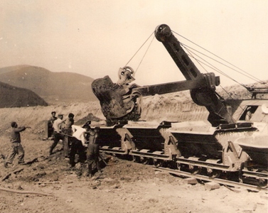  Lyžica lanového bagra nakladá plechové huntíky. Asistujú robotníci v pracovnom. V pozadí kopce. Text: Stavba trate Turňa - Rožňava. Anon., cca 1950. 150 x 123 