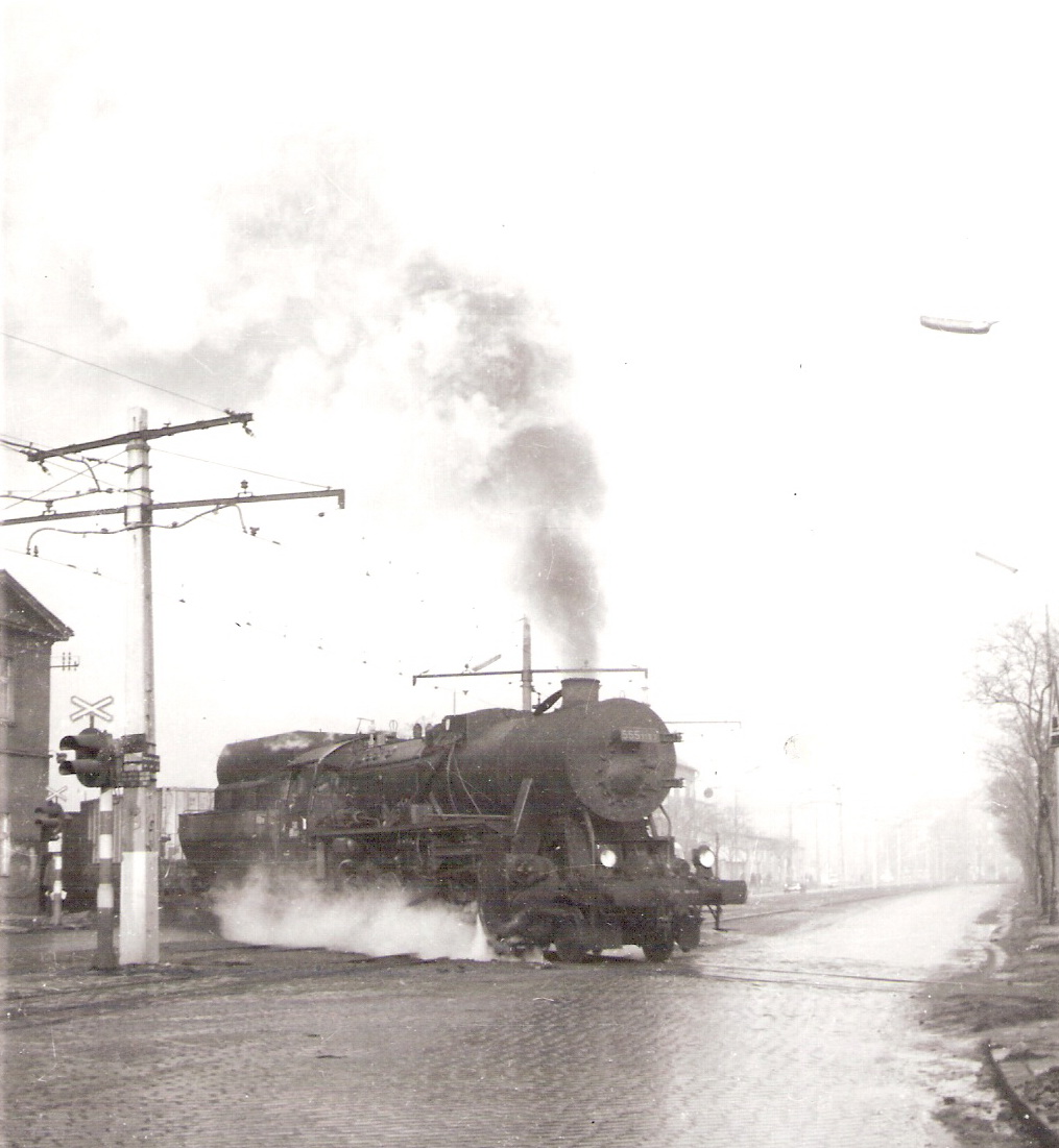  Parný rušeň 555.3163 na priec. Steinerova ul. (žst. Brat. filiálka - Bratislava Nivy). Pohľad zprava zpredu. Priecest. zab. zar, trolej električky, roh budovy koňky. Foto: M. Peciar, cca 1972. 130 x 178 