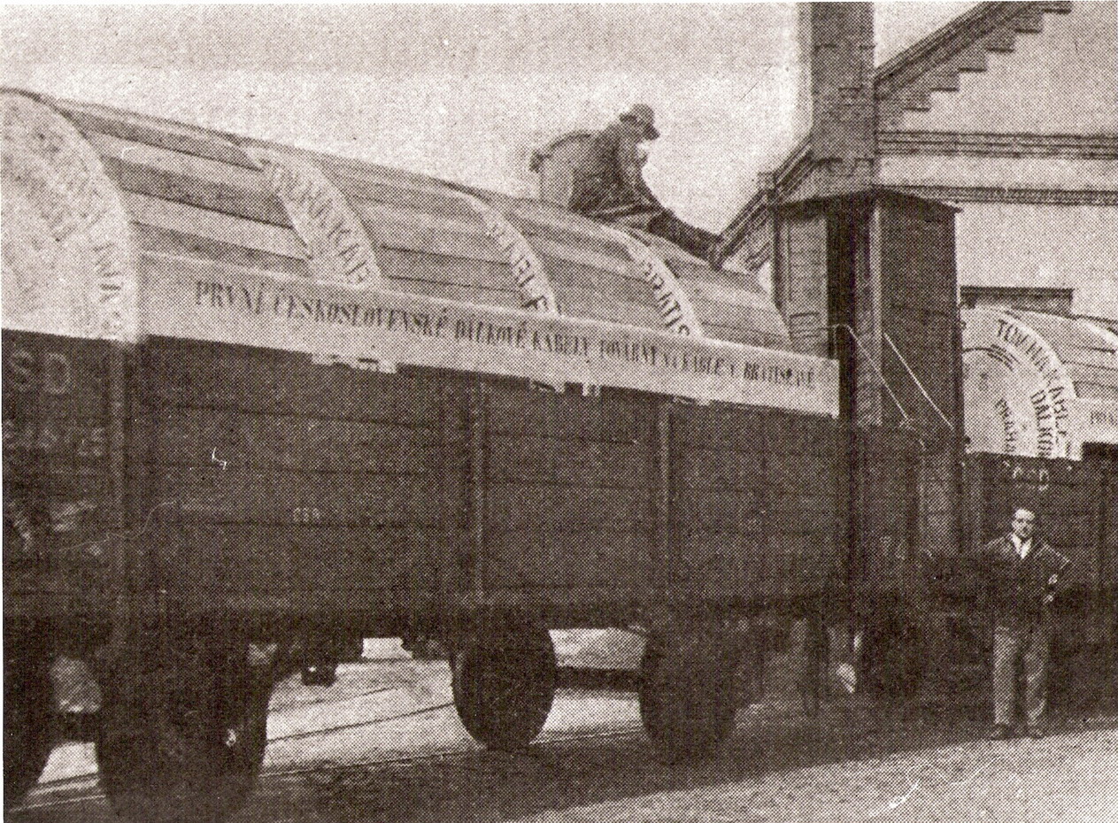  Nákladný dvojnápravový vysokostenný vozeň ložený zadebnenými kábelmi s nápisom TOV NA KABLY - PRVNÍ ČS DÁLKOVÉ KABELY... Pohľad predobočný. Vzadu priemysel. budova Továrne na káble v Bratislave. Foto: Anon, cca 1928. 229 x 169 