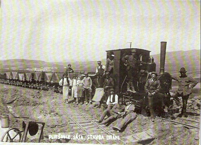  Úzkorozchodný parný rušeň s drev. huntíkmi. Pred ním skupina mužova a žien v civile i krojoch. Vľavo vpredu prenosná poľná vyhňa. Text: Výstavba trate Zvolen - Krupina. Pliešovce-Sása, stavba dráhy. Foto: Anon., cca 1923, farebná xerokópia. 112 x 83 