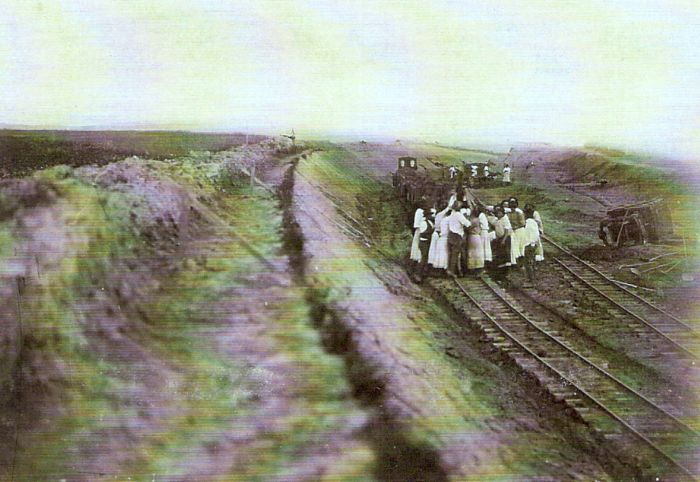  Široký plytký zárez, v ňom pracovná drážka s drev. huntíkmy, na vzdialenom konci s parným rušňom. Skupina mužov v krojoch a civile při práci. Text: Výstavba trate Zvolen - Krupina. Foto: Anon., cca 1923, farebná xerokópia. 128 x 90 