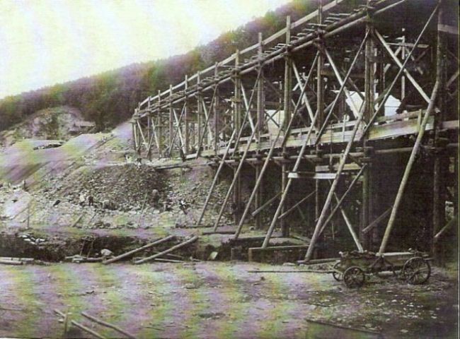  Veľké drevené lešenie so skružami pre betónovanie mostných oblúkov. Zalesnené úbočie, dole drevený konský povoz. Text: Výstavba trate Zvolen - Krupina. Foto: Anon., cca 1923, farebná xerokópia. 111 x 86 