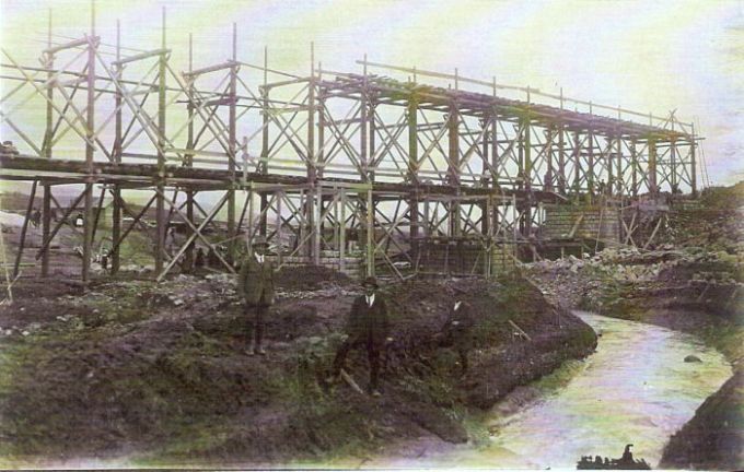  Veľké drevené lešenie, mostné piliere v stavbe. Vpredu traja muži v civile. Široký potok. Text: Výstavba trate Zvolen - Krupina. Foto: Anon., cca 1923, farebná xerokópia. 144 x 91 