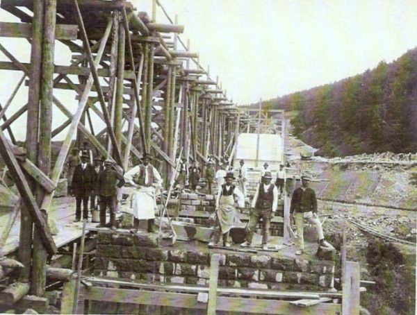  Veľké drevené lešenie, rozostavané piliere, muži v civile a pracovnom. Pohľad v osi lešenia. Vzadu zalesnený svah. ext: Výstavba trate Zvolen - Krupina. Foto: Anon., cca 1923, farebná xerokópia. 112 x 85 