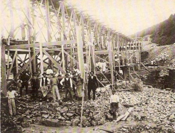  Veľké drevené lešenie, rozostavané pilier. Skupina mužov v pracovnom, civile a krojoch pózuje dole. Vzadu zalesnený svah. Text: Výstavba trate Zvolen - Krupina. Foto: Anon., cca 1923, farebná xerokópia. 113 x 88 