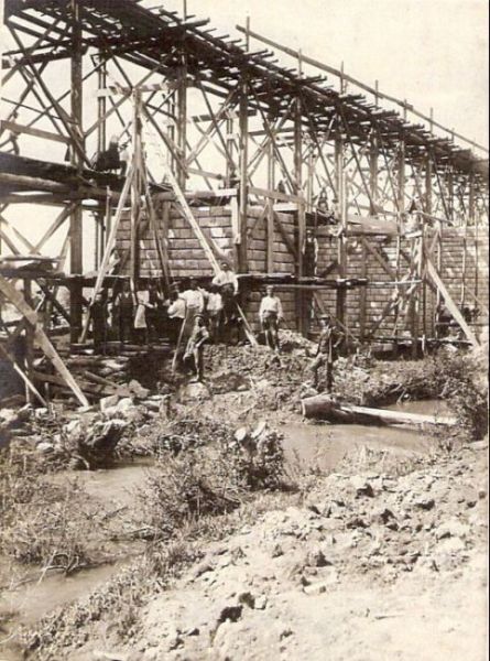  Veľké drevené lešenie, dole rozostavané piliere. Muži v civile a parcovnom. Text: Výstavba trate Zvolen - Krupina. Foto: Anon., cca 1923, farebná xerokópia. 84 x 112 