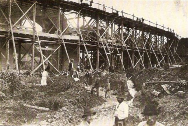  Veľké drevené lešenie, piliere a klenby v stavbe, vľavo dohotovená klenba. Dole potok. Pracuvúci muži v civile. Mechanický vrátok. Text: Výstavba trate Zvolen - Krupina. Foto: Anon., cca 1923, farebná xerokópia. 142 x 91 