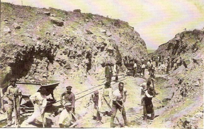  Hlinito-skalný zárez s poľnou drážkou a plechovými huntíkmi. Ľudia pri odkopávaní svahu. Text: Výstavba trate Zvolen - Krupina. Foto: Anon., cca 1923, farebná xerokópia. 144 x 90 