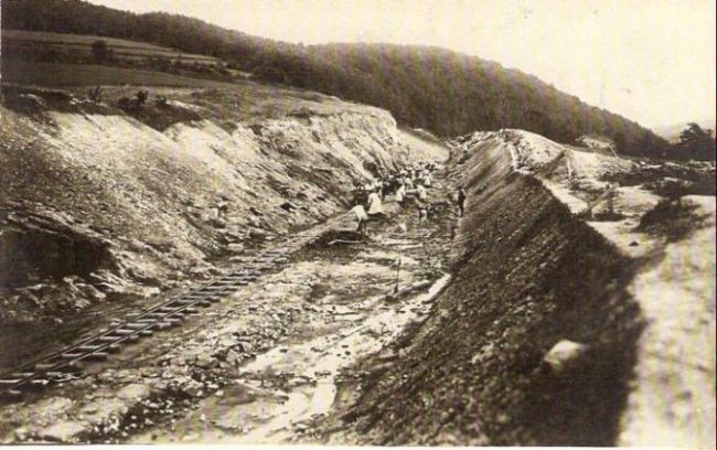  Plytký zárez s pracovnou drážkou. V záreze ľudia nakladajú plechové huntíky. Text: Výstavba trate Zvolen - Krupina. Foto: Anon., cca 1923, farebná xerokópia. 143 x 91 