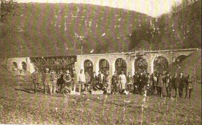  Dlhý most s radom kamenných oblúkov a oceľovým stredným trapézovým poľom s hornou mostovkou. Vpredu skupina civilov, vzadu hora. Text: Výstavba trate Zvolen - Krupina. Foto: Anon., cca 1923, farebná xerokópia. 144 x 90 