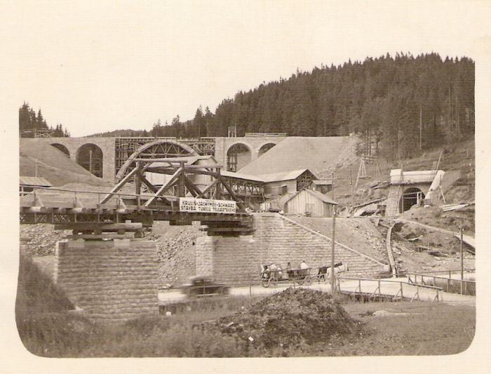  Stavenisko pri zastávke Telgárt-penzión počas výstavby trate. Pohľad zdola na rozostavaný most pred zastávkou, ústie tunela a viadukt nad zastávkou. Konečná stanica stavebnej lanovky, cesta s povozom a autom. Foto: Anon., cca 1933. 104 x 70 