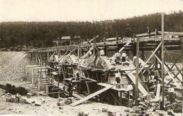  Rozostavaný veľký kamenný most. Rozpracované klenby, drevené lešenie. Ľudia pri práci. Vzadu násyp a les. Text: Výstavba trate Zvolen - Krupina. Foto: Anon., cca 1923, farebná xerokópia. 143 x 90 