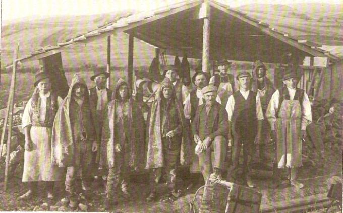  Skupina ľudí v civile a kroji, niektorí s kapucňami, pod dreveným prístreškom. Vzadu horská krajina. Text: Výstavba trate Zvolen - Krupina. Foto: Anon., cca 1923, farebná xerokópia. 145 x 90 