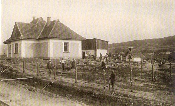  Malá výpravná budova, kôlňa a chlebová pec. Vpredu kôly pre plot záhrady. Pózujúci ľudia v civile a krojoch. Vzadu obec a horské úbočie. Text: Výstavba trate Zvolen - Krupina. Foto: Anon., cca 1923, farebná xerokópia. 140 x 90 