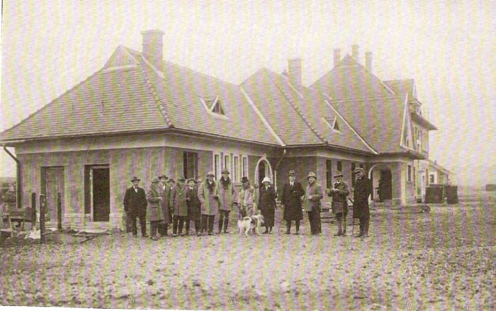  Výpravná budova - nárožný pohľad z predstaničného priestoru. Pred domom skupina civilov. Text: Výstavba trate Zvolen - Krupina. Foto: Anon., cca 1923, farebná xerokópia. 143 x 92 