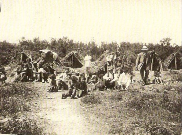  Ľudia - muži, ženy i deti - sediace a postávajúce na zarastenom svahu s vyšliapanou cestou. Vzadu prístrešky z čečiny. Text: Výstavba trate Zvolen - Krupina. Foto: Anon., cca 1923, farebná xerokópia. 110 x 83 