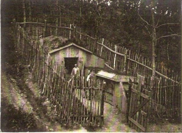 Oplotený drevený prístrešok v zalesnenom úbočí, v ňom dva stojaci muži. Text: Výstavba trate Zvolen - Krupina. Foto: Anon., cca 1923, farebná xerokópia. 110 x 83 