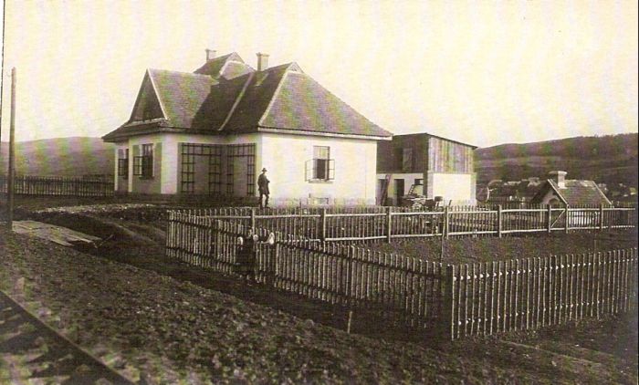  Malá výpravná budova, kôlňa a chlebová pec. Vpredu oplotená záhrada. Pózujúci muž a žena. Vľavo trať a telegraf. tyč. Vzadu obec a horské úbočie. Text: Výstavba trate Zvolen - Krupina. Foto: Anon., cca 1923, farebná xerokópia. 143 x 90 