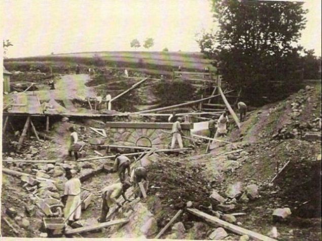  Kamenný železničný priepust v stavbe. Ľudia pri murárskych prácach na odvádzacom kanále. Nezarastený svah s prašnou cestou, drevené lávky a lešenia. Text: Výstavba trate Zvolen - Krupina. Foto: Anon., cca 1923, farebná xerokópia. 112 x 83 