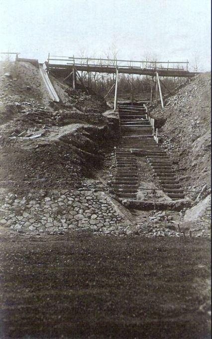  Provizórny drevený mostík nad schodovito usporiadanou sklonenou plochou. Vzadu stromy bez listov. Text: Výstavba trate Zvolen - Krupina. Foto: Anon., cca 1923, farebná xerokópia. 90 x 140 