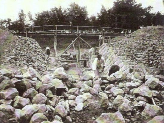  Kamenný priepust v práci. Rozostavané opory, drevené provizórne premostenie. Dole kopa kameňa, ľudia pri práci. Vzadu stromy. Text: Výstavba trate Zvolen - Krupina. Foto: Anon., cca 1923, farebná xerokópia. 110 x 83 