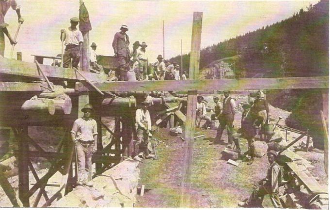  Železničný most v stavbe. Pohľad v osi koľaje. Vľavo proviz. drev. mostovka a stav. drážka, ľudia v civile a krojoch. Vzadu zalesnený svah, dole odkopaný. Text: Výstavba trate Zvolen - Krupina. Foto: Anon., cca 1923, farebná xerokópia. 144 x 92 