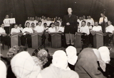  Pohľad cez hlavy poslucháčov na konferenciéra a estrádny orchester so zmiešaným zborom. Text: Trať Družby - Spišské Vlachy. Anon., cca 1950. 175 x 122 