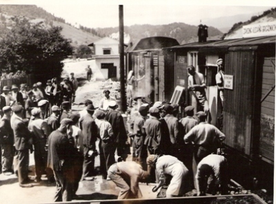  Skupina ľudí v pracovnom čakajúcich na vydaj stravy z krytého vozňa s nápisom Závodná kuchyňa brigádnikov. Text: Trať Mládeže. Anon., cca 1950. 163 x 120 