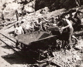  Skupina mladých robotníkov (jeden s vojenskou lodičkou) pri nakladaní plech. huntíku kameňmi. Text: Trať Mládeže, výstavba trate. Anon., cca 1950. 142 x 115 