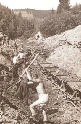  Skupina robotníkov pri ručných zemných prácach v hlinitom záreze. Pracovná drážka, plech. huntíky. Text: Trať Družby. Foto: ČSD, cca 1950. 125 x 82 