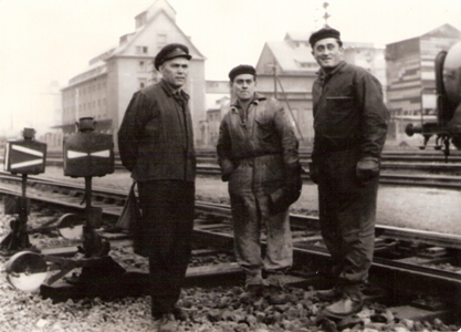  Traja muži v prac. (jeden v železn. čiapke a s práporkom) stojaci pri styku dvoch per. výhybiek a 8-bokými vým. lamp. Za nimi priemyselné budovy. Anon., cca 1960. 180x130 