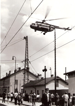  Rekonštrukcia trakč. vedenia v Spišskej Novej Vsi. Vrtulník s podves. lanom, robotník na trakč. podpere, výpr. budova, prizerajúci sa ľudia. Foto: M. Meliška, cca 1977. 122 x 175 