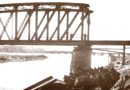  Pohľad zboku na poloparabolic. nosník mosta cez Váh pri Trenčíne (prvý od Zlatoviec) s chýbajúcou časťou oblúka a prísl. priečok. Torzo je podoprené dreveným pilierom. Vpredu mostné trosky. Foto: Jelínek, R., 1945. 129 x 90 
