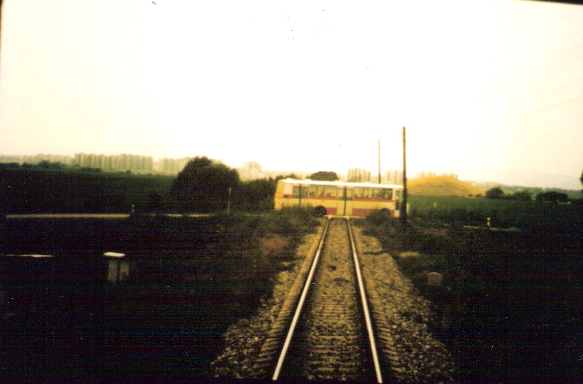  Trať Bratislava-Petržalka - Rusovce. Pohľad zo stred. dverí posled. vozňa na priamy úsek s telegraf. tyčami. Priecestie cesty do Jaroviec s autobusom. Foto: M. Entner, 1990. 155 x 100, COLOR 
