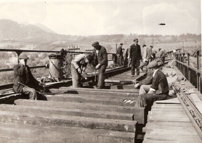  Poklád. zvršku na moste - teslov. mostníc, vz. práca s vrtulami. Zábradlie mosta a zrejme pec na nity. Ľudia v žel. rovnoš. i civile. Vz. hory. Text: Žel.zam. na opr. trate Handlová - H. Štubňa. Anon., cca 1945. 172 x 122 