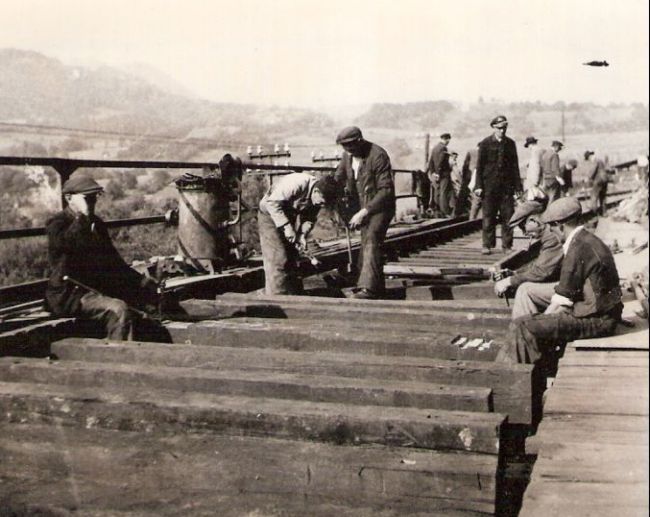  Pokládka zvršku na moste - teslov. mostníc, vz. práca s vrtulami. Zábradlie mosta a zrejme pec na nity. Ľudia v žel. rovnoš. i civile. Vz. hory. Text: Žel. zam. na opr. trate Handlová - H. Štubňa. Anon., cca 1945. 100 x 80 