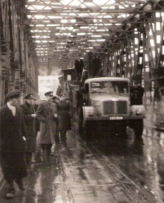  Prepr. ozdob. rušňa 310.4/5 (?) na podv. Cullmayer za Tatrou T 111 na bratisl. moste. Asfalt s dvojicou električk. koľají, ľudia vo voj. rovnoš. i prac. Text: Povojn. obnova prev. v okolí Petržalky. Anon., cca 1945. 117 x 138 