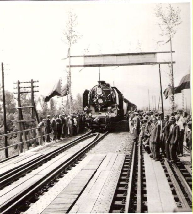  Ozdob.387.0 so znak.ČSR a päťroč.a s os.vlak. vchádza špalier. ľudí popod slavobránu na ozdob.dvojkoľ. most. Telegr. tyče. Text: Sprevádzk. mosta cez Váh pri Šali po 2. svet. vojne. Anon., cca 1950. 124 x 135 