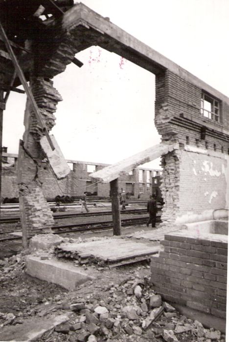  Pohľad otvorom vybúraným v tehlovom múre budovy s betón. vencom bez strechy do interiéru s koľajami. Vpredu tehlová suť a múrik. Text: Dielne Prešov. Anon., cca 1945. 114 x 170 