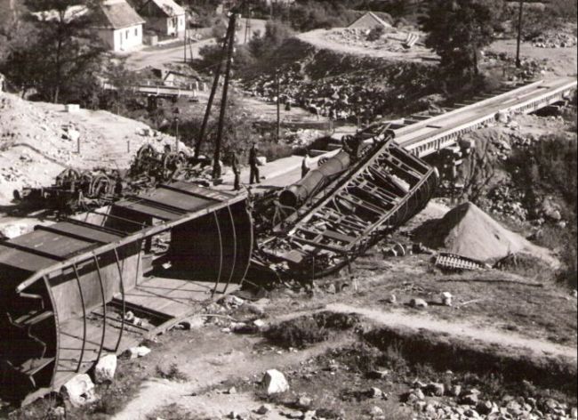  Z. Podkriváň. Prevrát. orabov. dvojnápr. nákl. a os. vozeň (plyn osvetl.). Trať na drev. most. provizóriu, pod ním vozň. spodok. Prích. cesta, plyn. lampa na oceľ stĺpiku. Dole obyt. domy a cesta. Anon., cca 1945. 173 x 125 