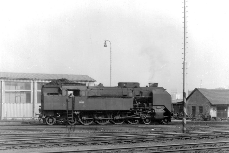  Parný rušeň 464.053 v žst. Klatovy. Pohľad zprava. Vzadu dielenské budovy, osvetľovacie stožiare a návestidlo AŽD. Anon., 7.4.1981. 140 x 95 